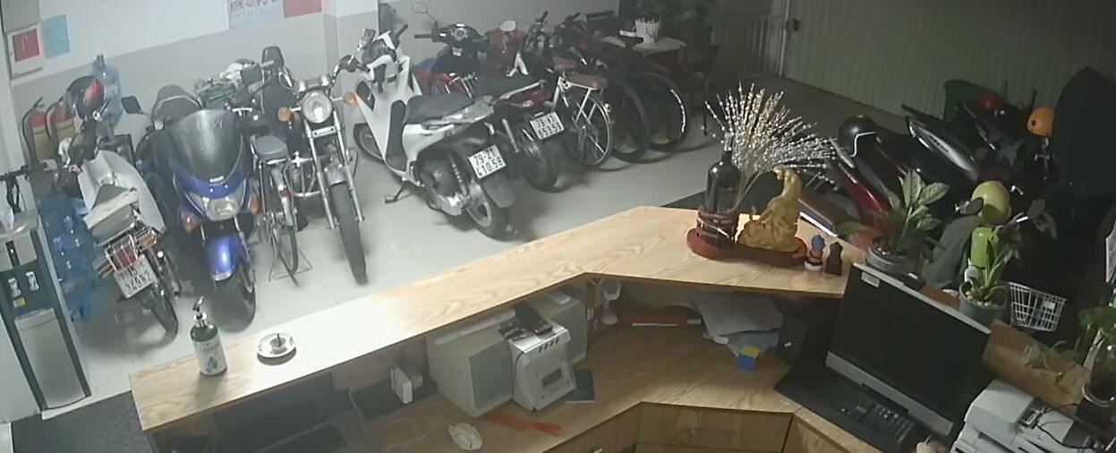 A room with a desk cluttered with various items, including a computer, a keyboard, and a small statue. There are multiple motorcycles parked in the background.