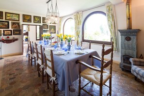 Ein Esszimmer mit einem langen Tisch, gedeckt für sechs Personen, umgeben von Holzstühlen. Ein großes Fenster mit Vorhängen, ein Steinplattenboden und ein alter Herd in der Ecke.