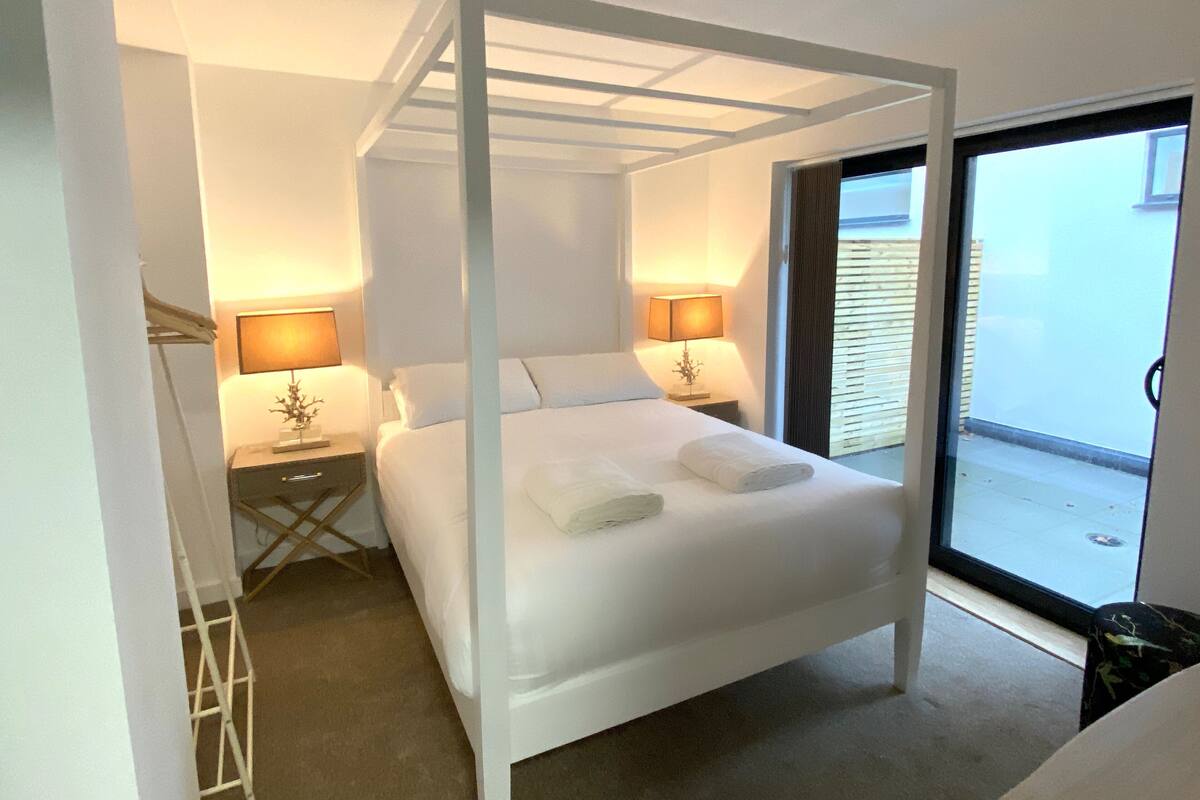 A modern bedroom with a four-poster bed, two bedside tables with lamps, and a view of the outside through a sliding glass door.