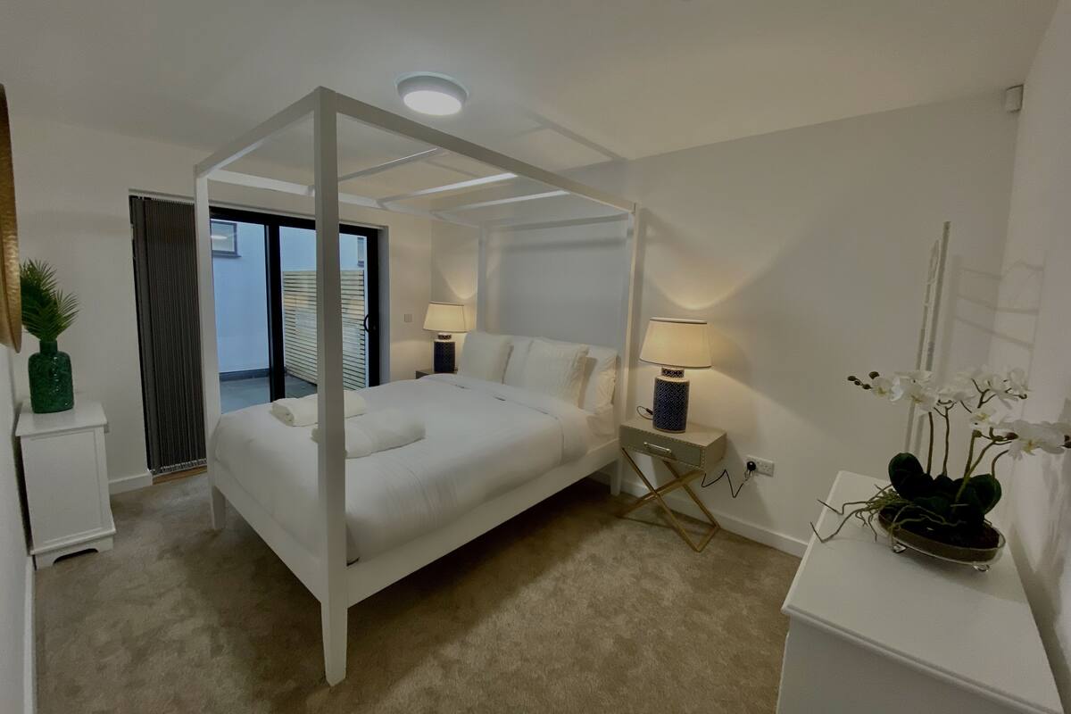A bedroom with a canopy bed, a nightstand with a lamp, a small potted plant, and a view of the outside through a sliding door.