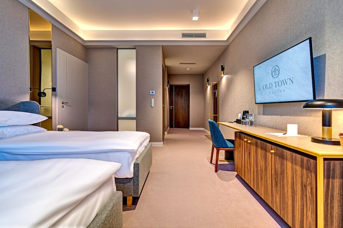 A hotel room with two beds, a wooden desk, a TV, and a hallway leading to another room.