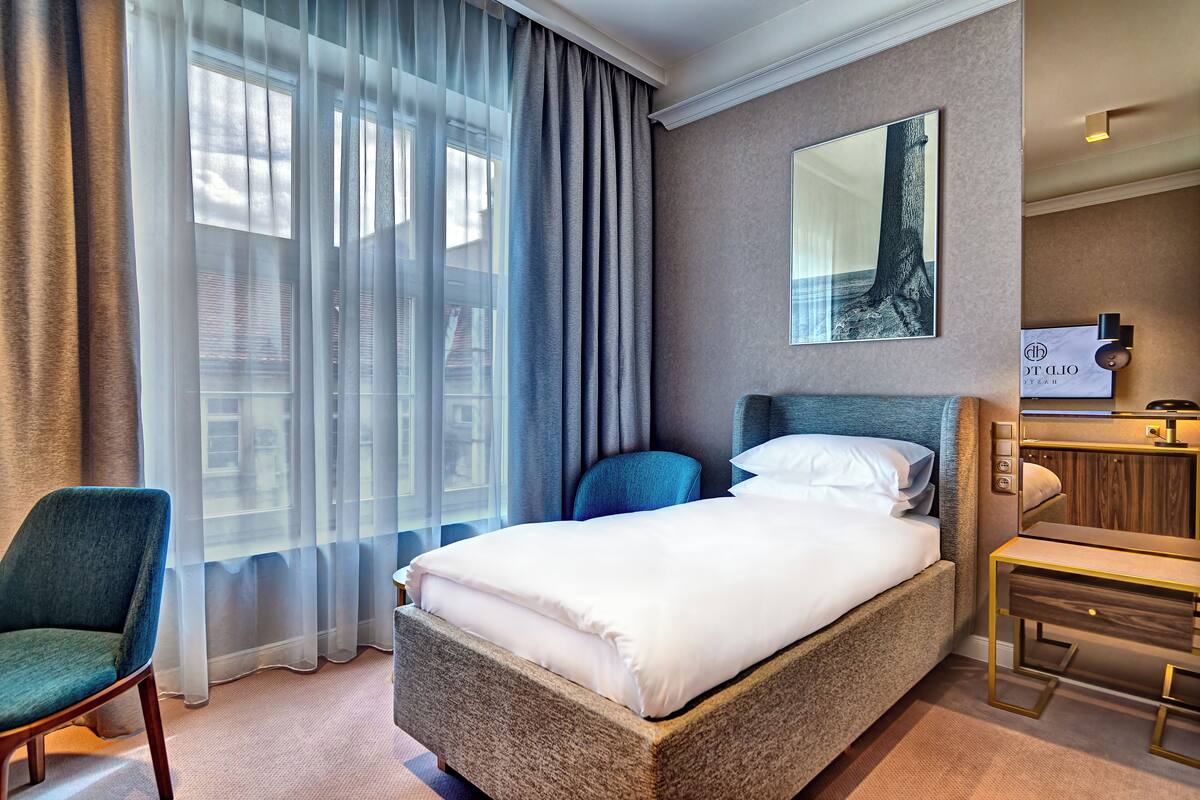 A hotel room with a large bed, a desk, a chair, and a window with sheer curtains.