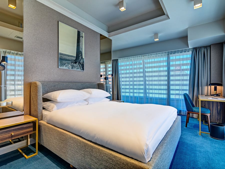 Quarto de hotel com uma cama grande, uma escrivaninha, uma cadeira e vista para a cidade através de amplas janelas.