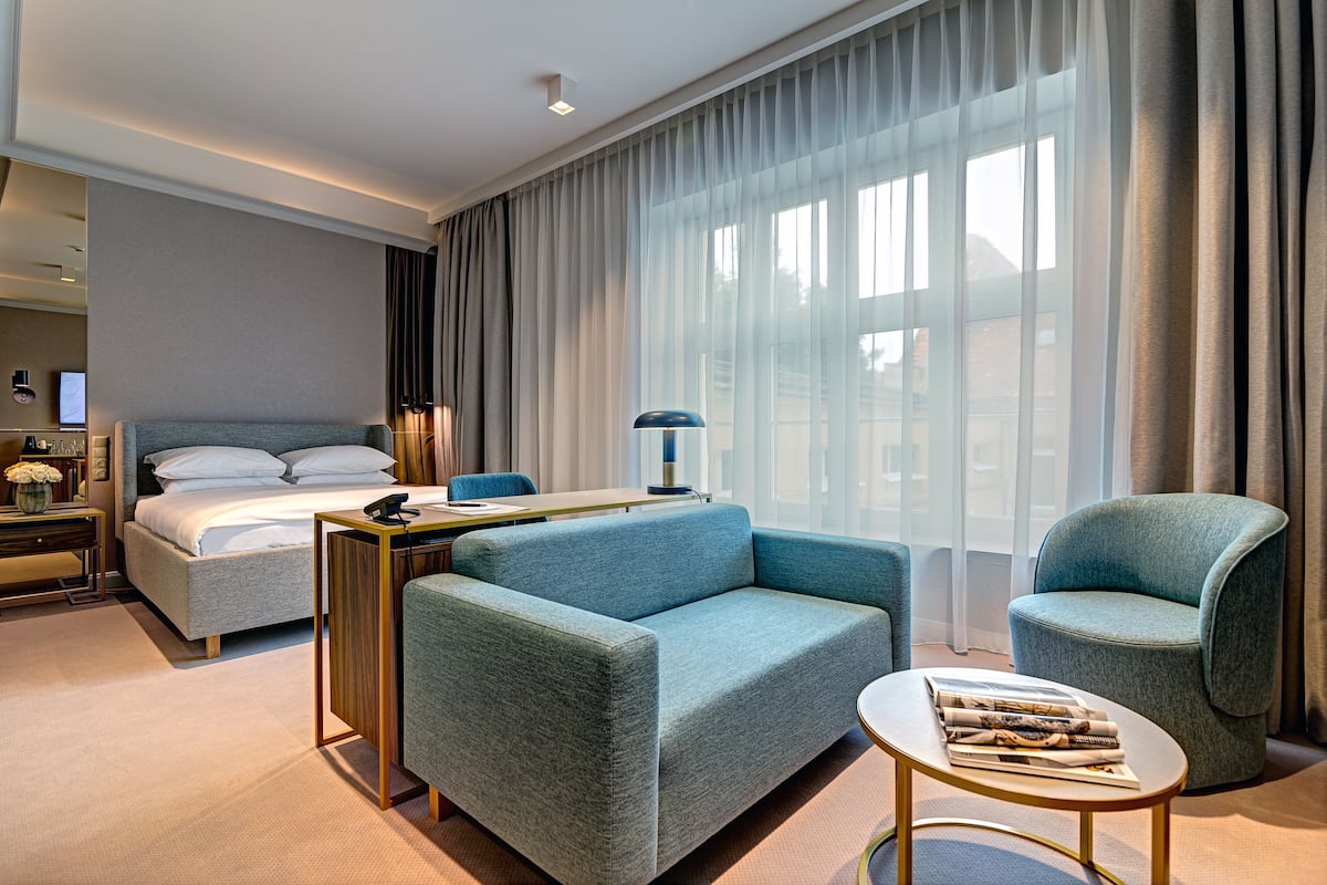 A modern hotel room with a bed, a sofa, a chair, a small table, and a large window with curtains.