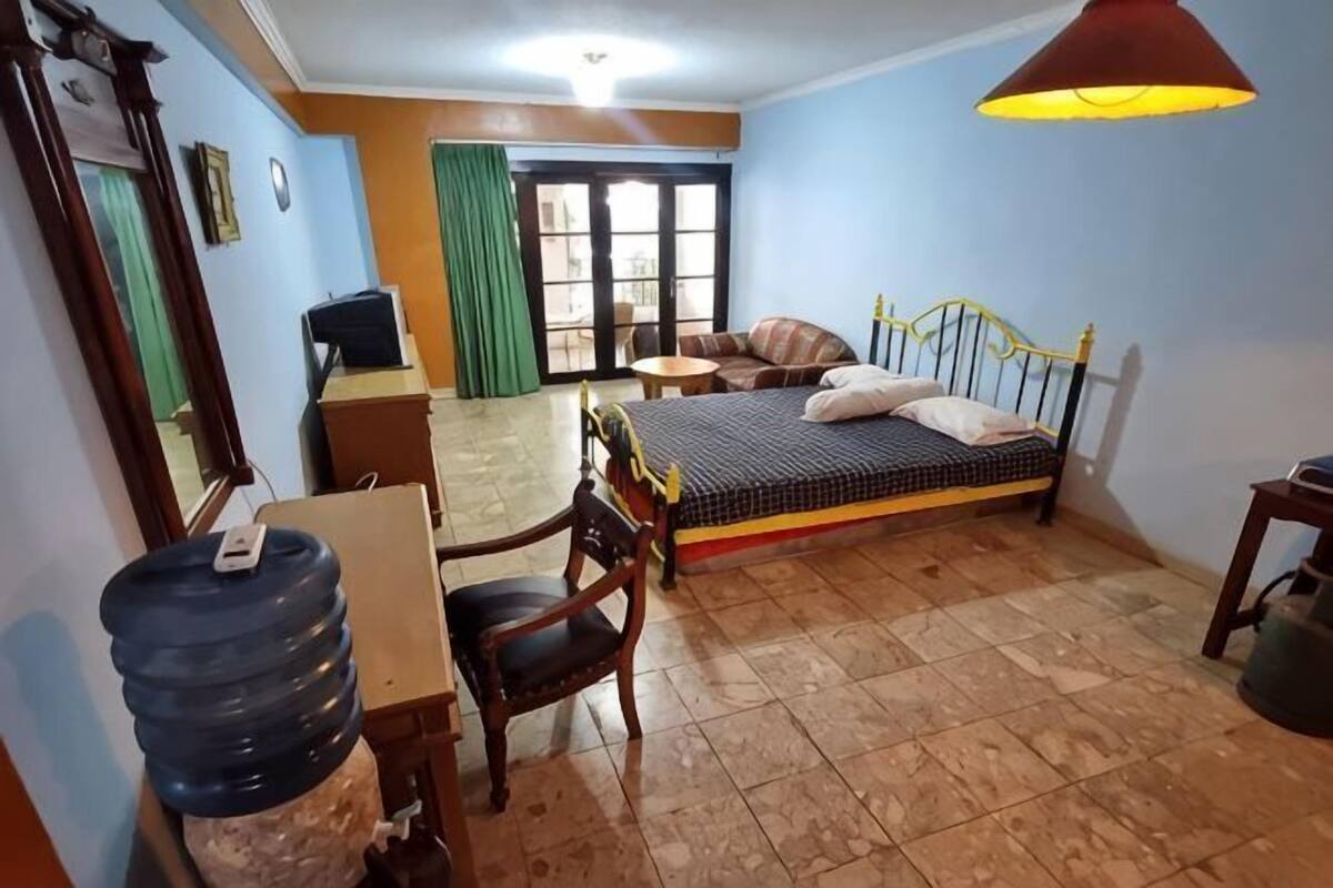 Une chambre à coucher comprenant un lit, un bureau, une chaise, un distributeur d’eau et une fenêtre avec des rideaux.