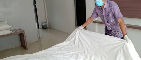 A person adjusting a white bedspread in a room with a wooden bench and a wall-mounted television.