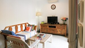 A living room with a sofa, a coffee table, a TV on a wooden cabinet, a wall clock, and a lamp. - Dewivilla Hotel (Padangbai)