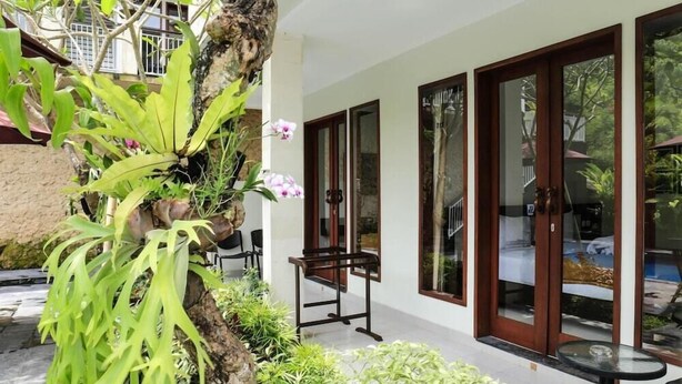 A modern building with large glass doors, a small outdoor seating area with a table and chairs, and lush greenery including a tree and various plants. - Dewivilla Hotel (Padangbai)