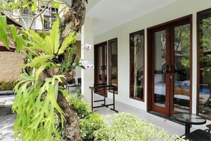 Un bâtiment moderne doté de grandes portes vitrées, d’un petit espace extérieur aménagé avec une table et des chaises, et d’une végétation luxuriante comprenant un arbre et diverses plantes.