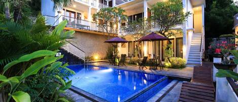 A swimming pool with a wooden deck, surrounded by lush greenery and a building with balconies.