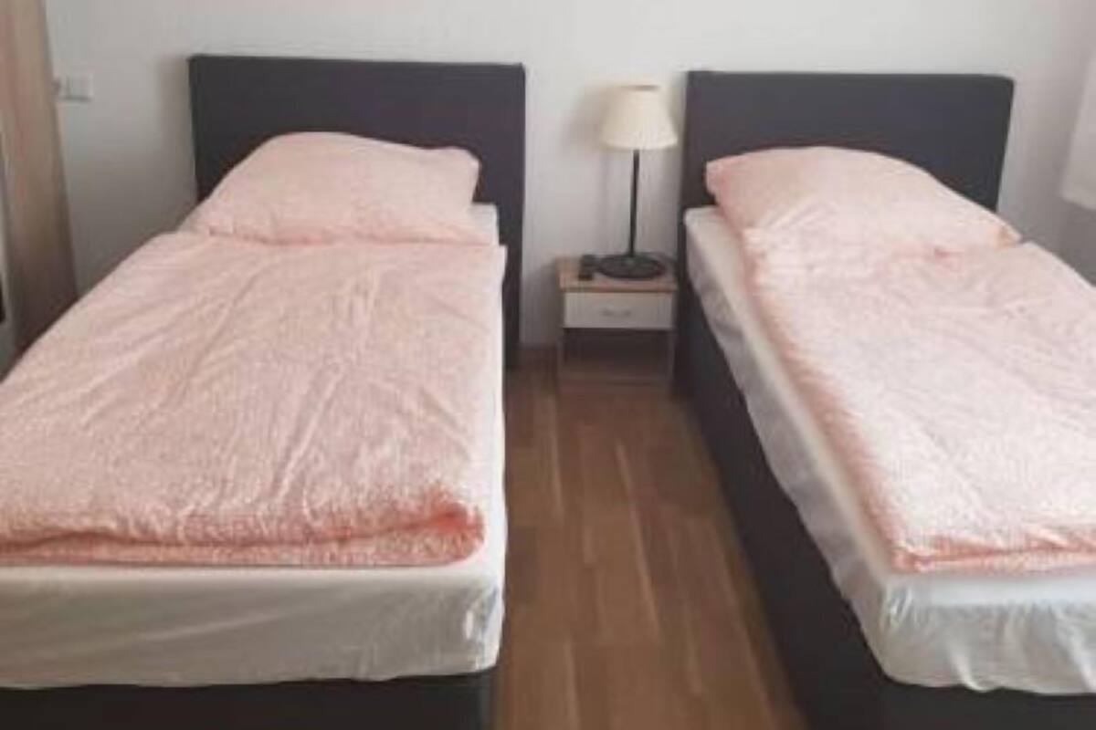 A room with two beds, each with a dark headboard and a pink blanket. There is a small bedside table with a lamp between the beds.
