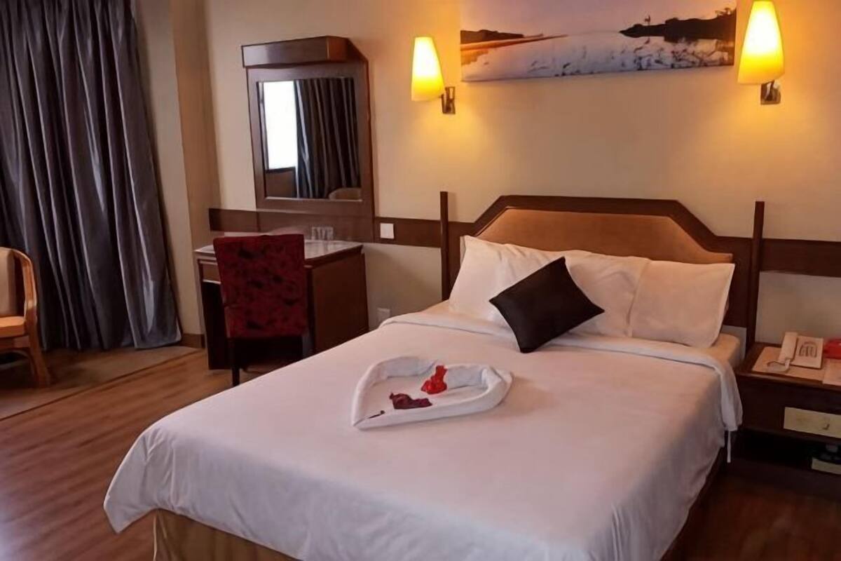 A hotel room with a bed, a desk, a chair, a nightstand, and a wall-mounted lamp.