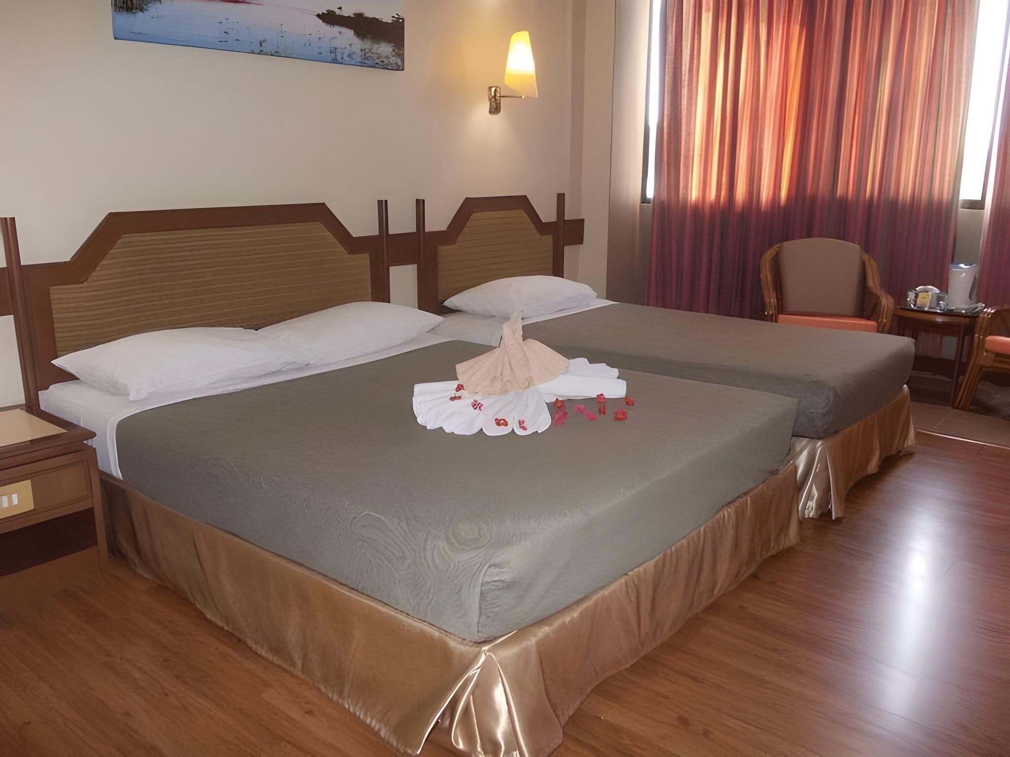 A hotel room with two beds, wooden headboards, a painting on the wall, a lamp, a chair, and a small table with a pitcher.