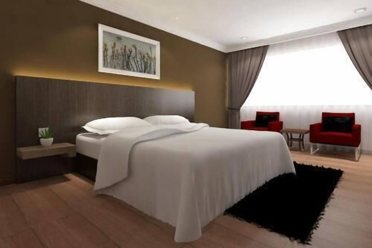 A modern hotel room with a large bed, a small table, two red chairs, a painting on the wall, and a large window with curtains.