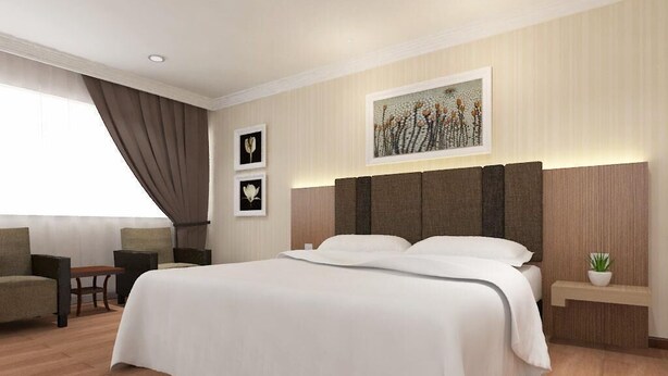 A hotel room with a large bed, a desk, a chair, a small table, a nightstand, and a potted plant. - Hotel Anika (Kluang)