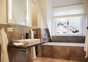 Une salle de bain moderne avec un lavabo, un miroir et une baignoire. Il y a une fenêtre qui donne sur une maison à l’extérieur.