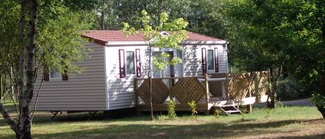 A small, single-story mobile home with a red roof, surrounded by trees and a wooden deck.