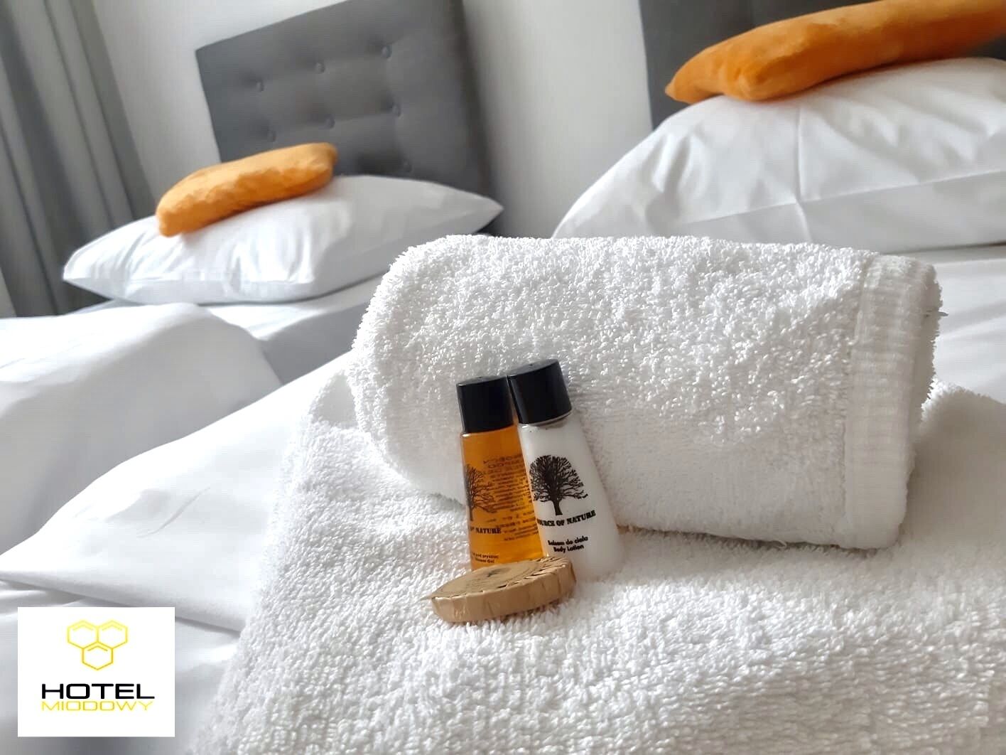 A neatly made bed with white linens, a folded white towel, and a bottle of body lotion on a wooden coaster.