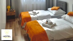 Une chambre d’hôtel avec deux lits, chacun recouvert d’une couvre-lit orange et agrémenté d’oreillers blancs, un téléviseur à écran plat fixé au mur, une table de chevet en bois avec une lampe et une bouteille, et une grande fenêtre avec des rideaux légers.
