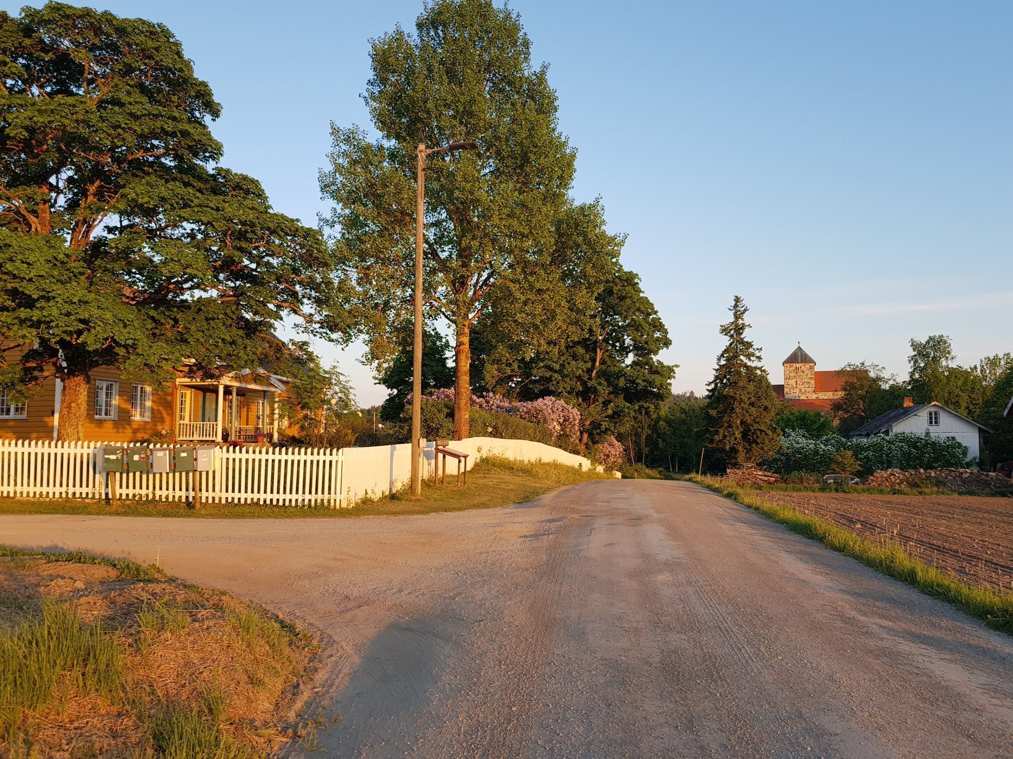 A rural road with a white picket fence, a house with a porch, and a stone tower in the background.