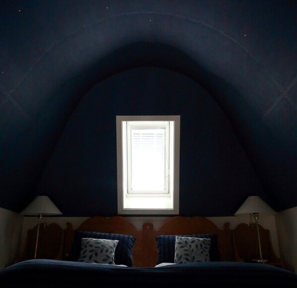Un dormitorio con techo inclinado, una ventana, una cama con almohadas y lámparas de noche.
