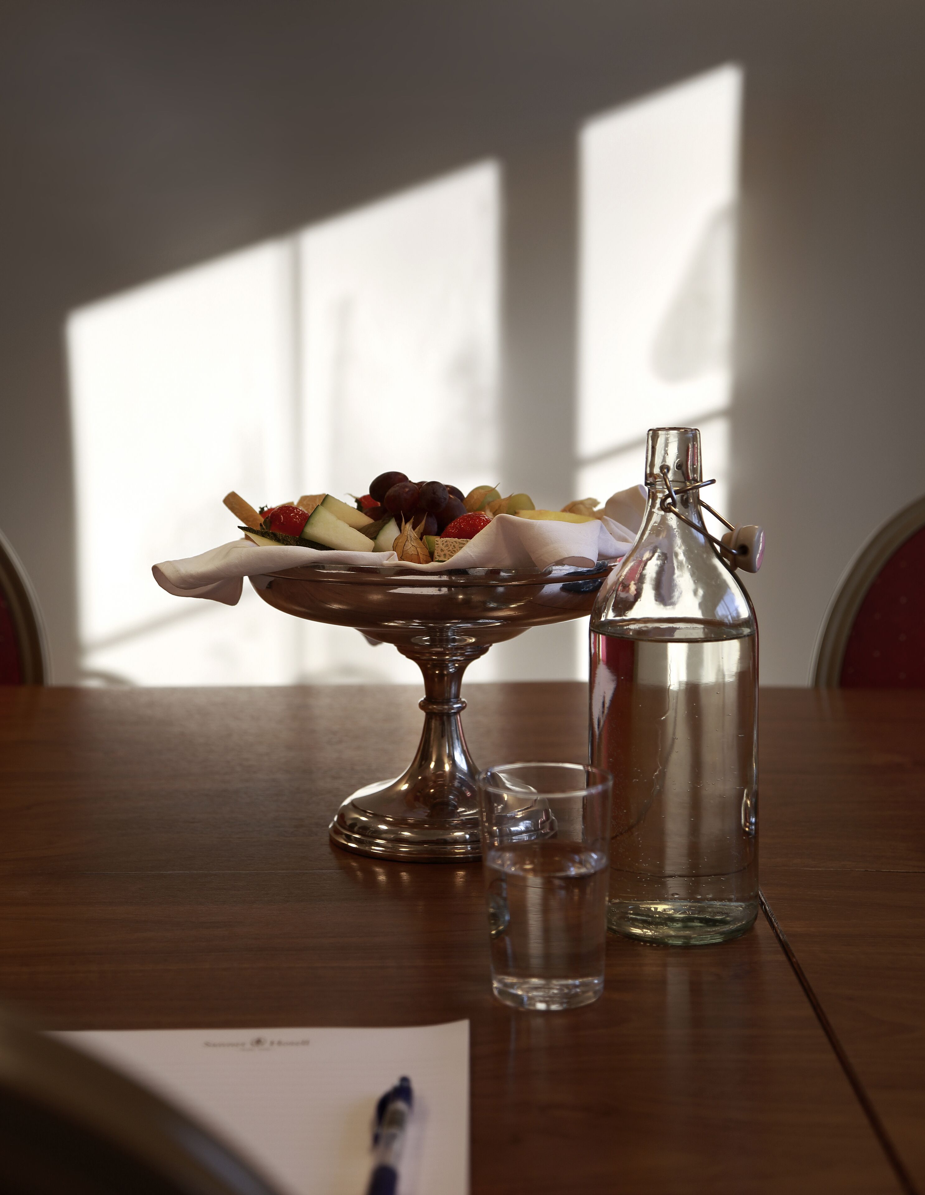 Uma mesa de madeira com uma fruteira, uma garrafa de vidro e um copo de água.