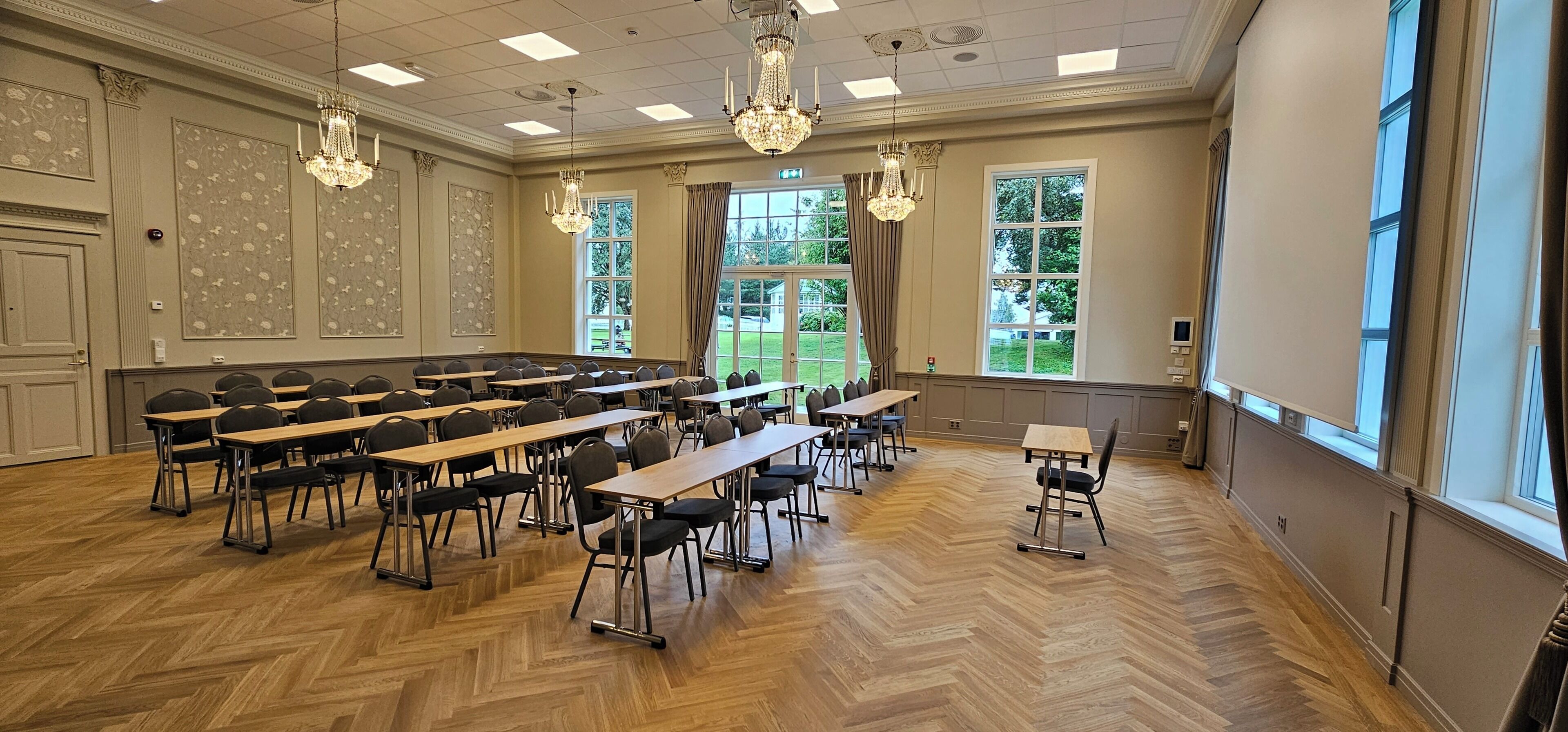 A conference room with multiple tables and chairs, large windows with curtains, and chandeliers.