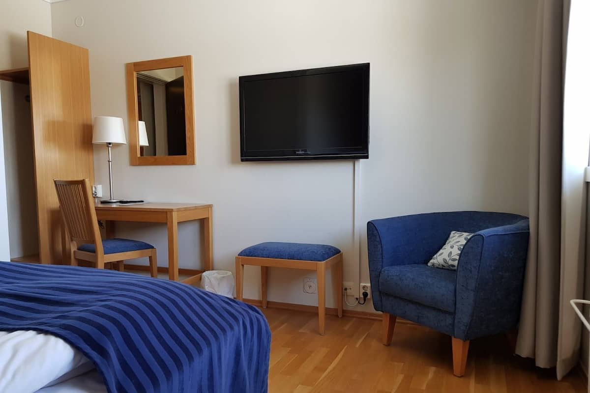 A hotel room with a bed, a desk with a chair, a television, and a blue armchair.