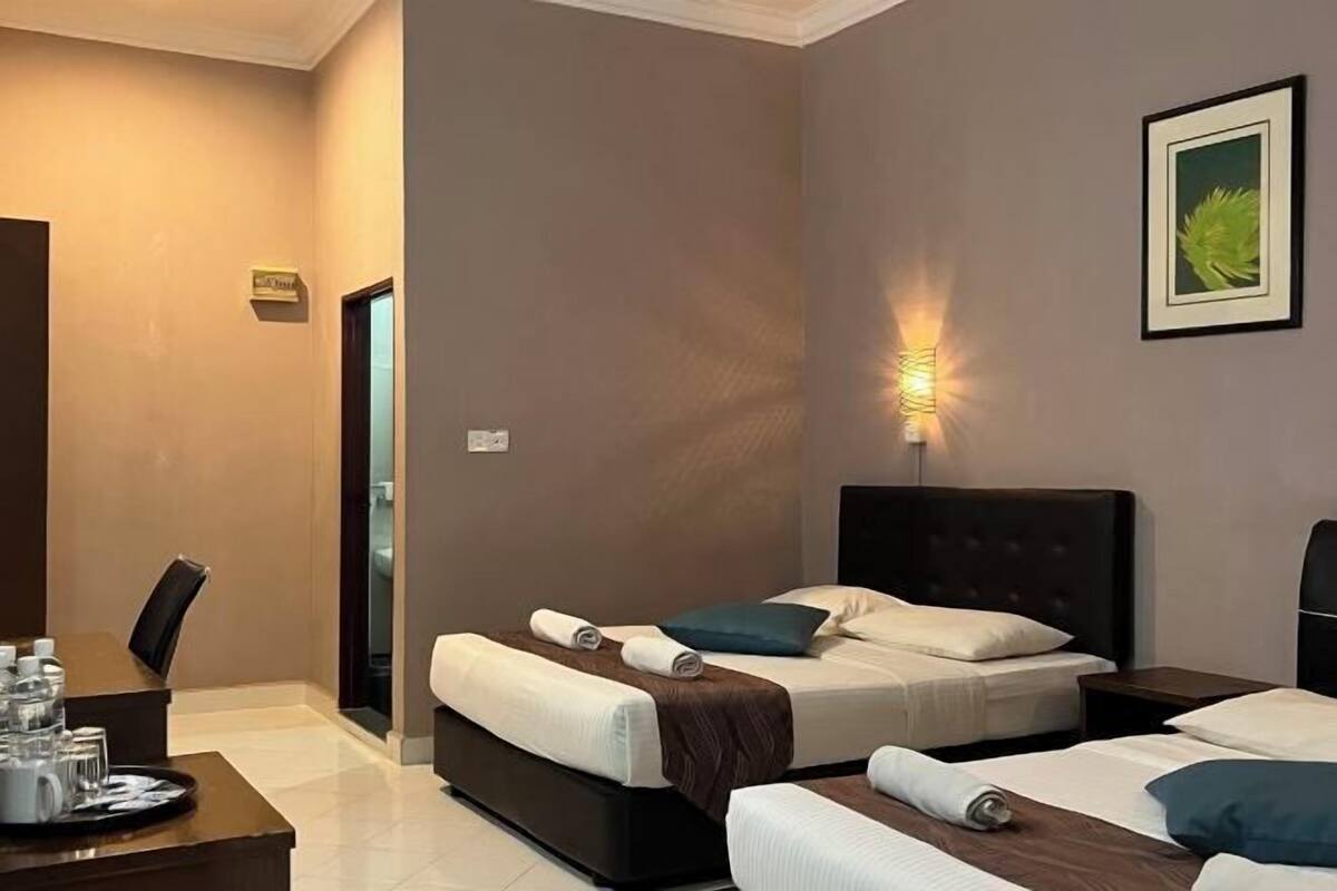A hotel room with two beds, a desk with a tray of glasses and a water bottle, and a wall-mounted light.