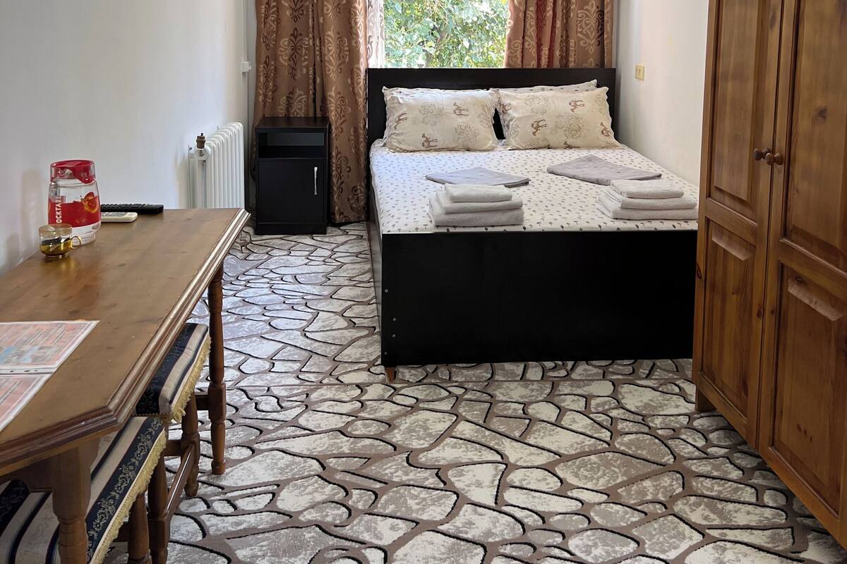 A bedroom with a bed, a wooden wardrobe, a table with a glass, a radiator, and a window with curtains.