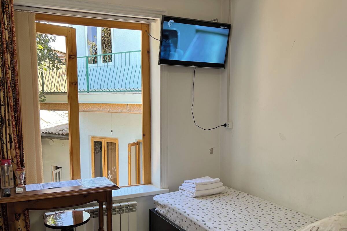 A small, tidy room with a single bed, a table with papers, a radiator, a TV mounted on the wall, and a window with a view of a building outside.