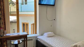 A small, tidy room with a single bed, a table with papers, a radiator, a TV mounted on the wall, and a window with a view of a building outside.
