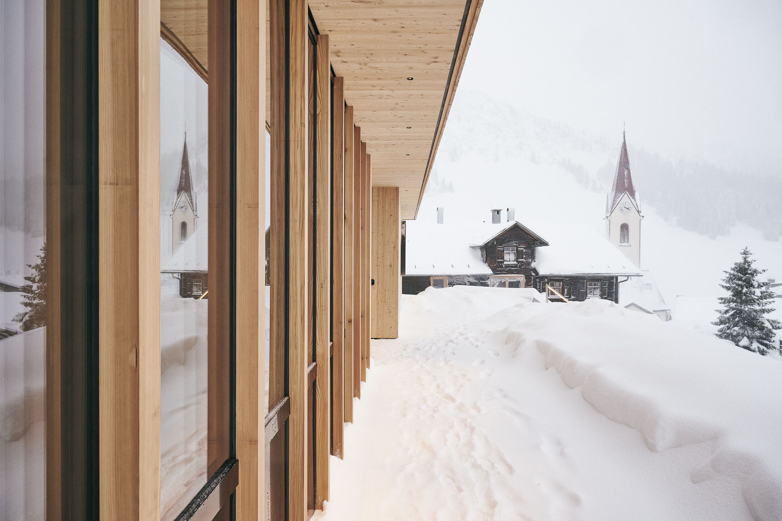 Un sentiero innevato che conduce a un edificio in legno con ampie finestre, con una chiesa sullo sfondo.