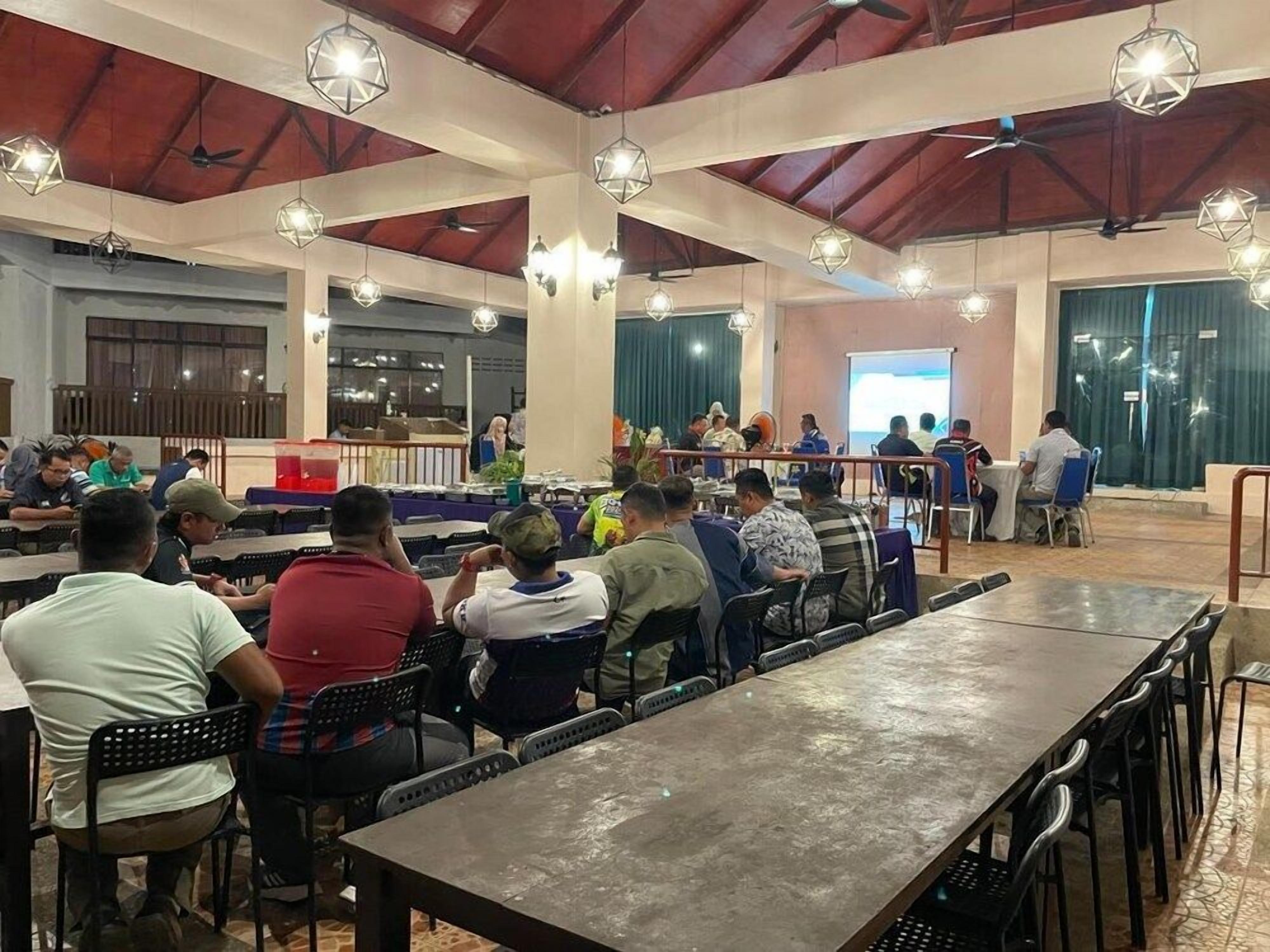 A spacious indoor dining area with multiple tables and chairs, a stage with a presentation screen, and hanging pendant lights.