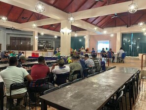 A spacious indoor dining area with multiple tables and chairs, a stage with a presentation screen, and hanging pendant lights. - Mersing Beach Resort (Mersing)