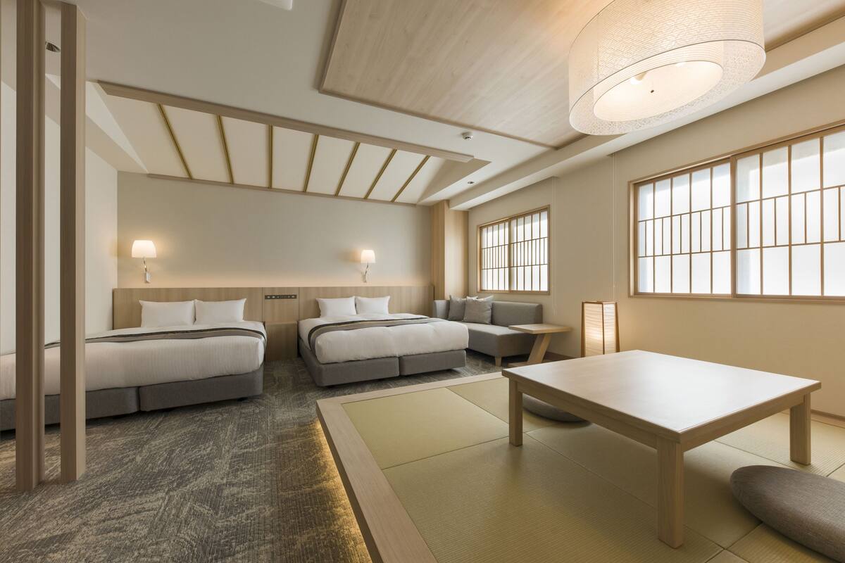A modern hotel room with two beds, a sofa, a table, and large windows.