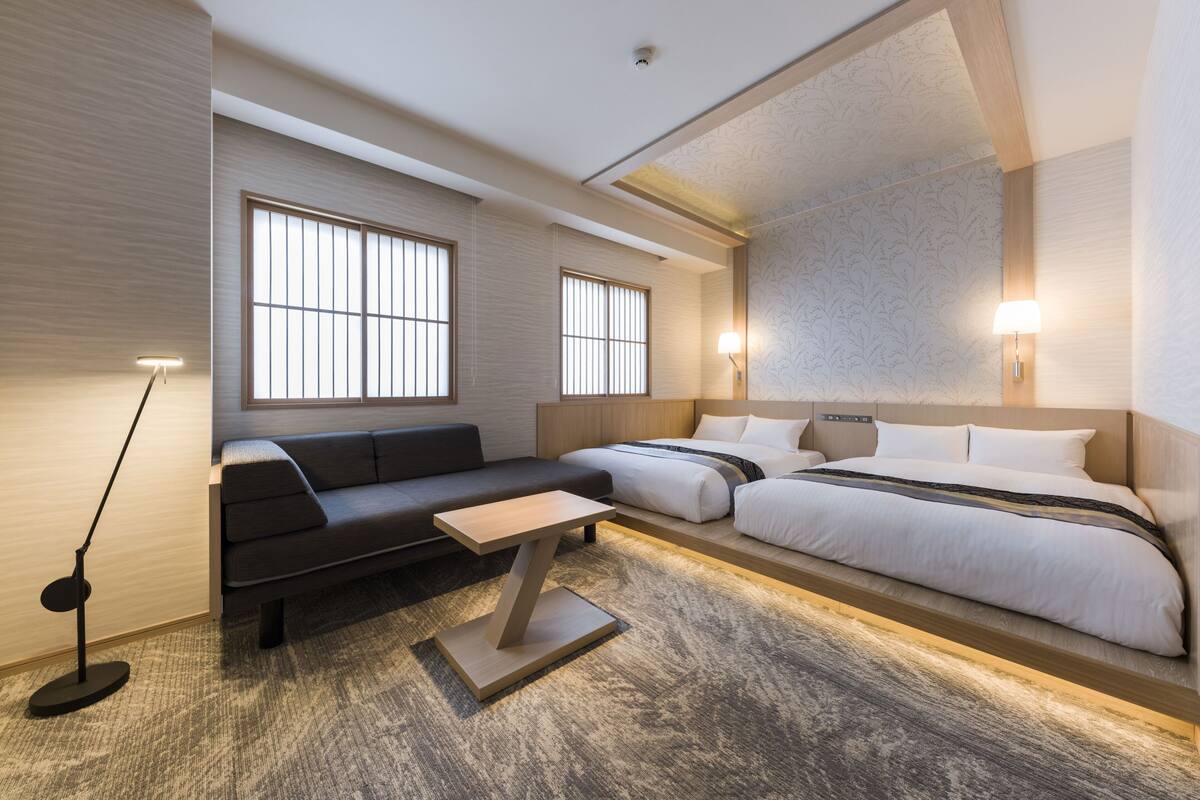 A modern hotel room with a sofa, two beds, a small table, and large windows.