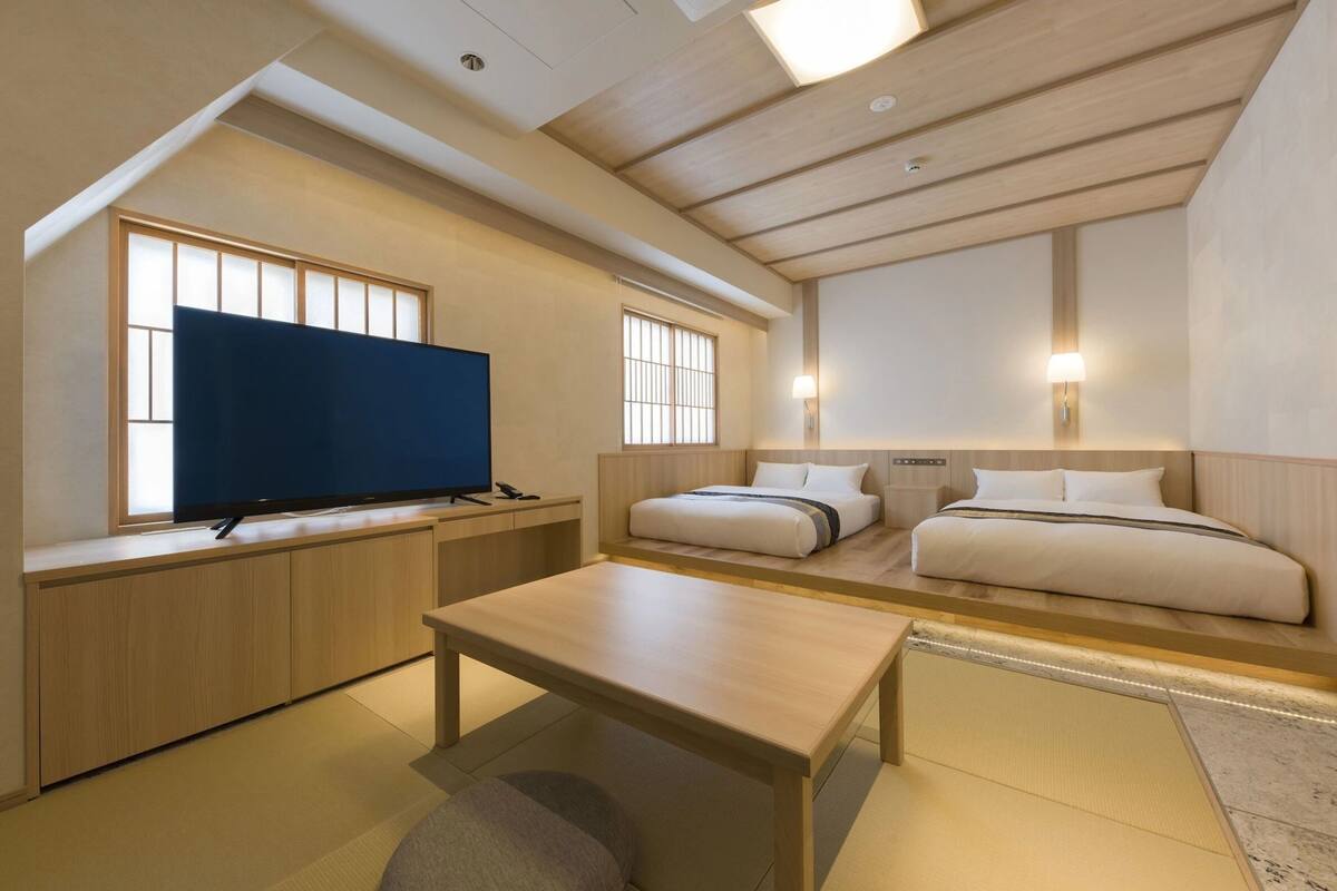 A modern hotel room with a flat-screen TV, a desk, two beds, and a large window with traditional-style blinds.