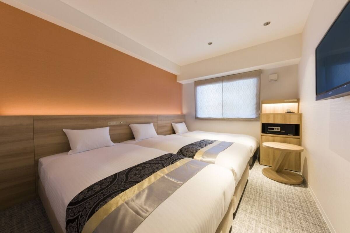 A hotel room with two beds, a TV, a small table, and a window with blinds.