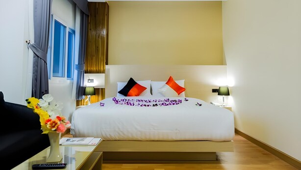 A hotel room with a bed, bedside lamps, a nightstand, a remote control, and a vase with flowers. - GT Residence Hotel (Buriram)