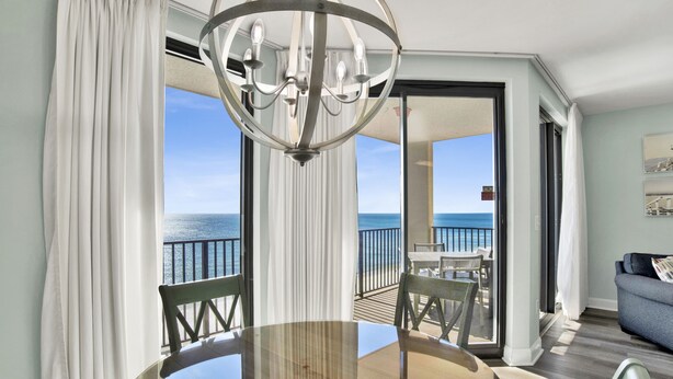 A dining area with a round table, chairs, and a chandelier. There is a balcony with a view of the ocean. - Beachfront 1BR @ Phoenix X • Sleeps 6 • Gulf Views, Pools & Next to Flora-Bama (Orange Beach)