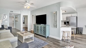 A modern living room with a sofa, coffee table, and TV, connected to a kitchen with a refrigerator and bar stools. - Beachfront 1BR @ Phoenix X • Sleeps 6 • Gulf Views, Pools & Next to Flora-Bama (Orange Beach)