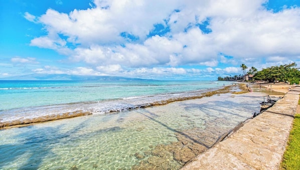 Una playa con aguas claras y poco profundas, un camino de piedra y algunos edificios al fondo.