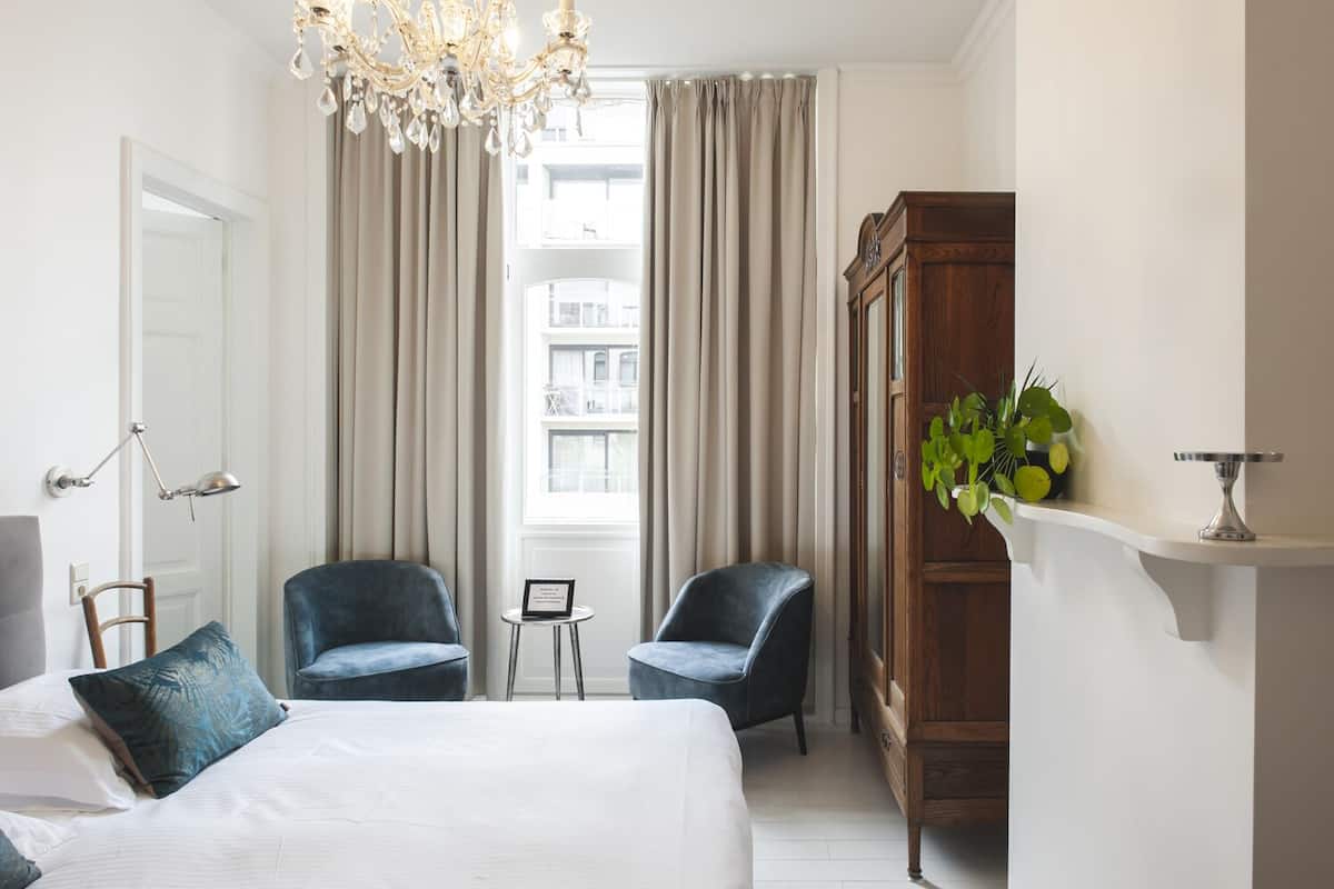Une chambre d’hôtel avec un grand lit, deux fauteuils, une armoire en bois, une petite table avec un panneau et une grande fenêtre avec des rideaux.