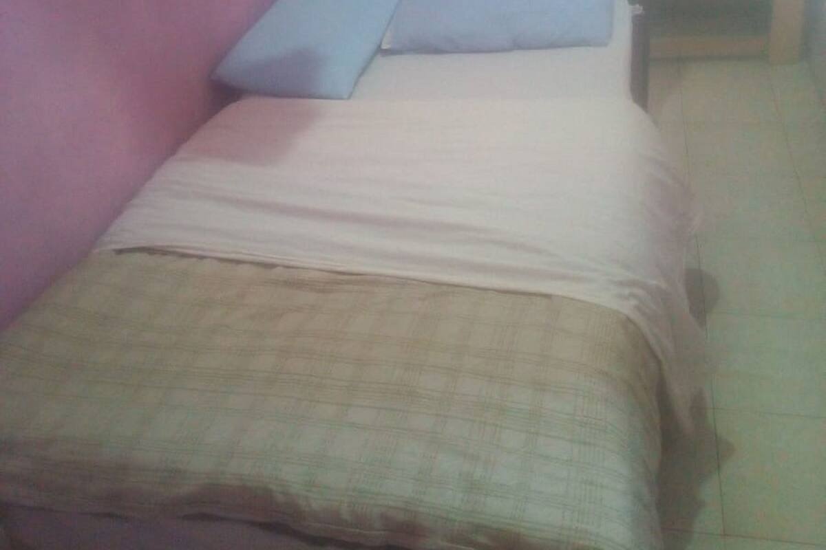 A small, single-bed room with a wooden headboard, a bedside table, and a wall-mounted hanging.