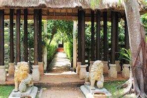 Stroll along the serene garden pathway to a charming thatched-roof pavilion, surrounded by lush vegetation and traditional stone statues.