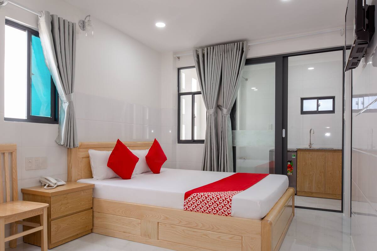 A modern hotel room with a wooden bed, a bedside table with a phone, a small desk, and a cabinet.