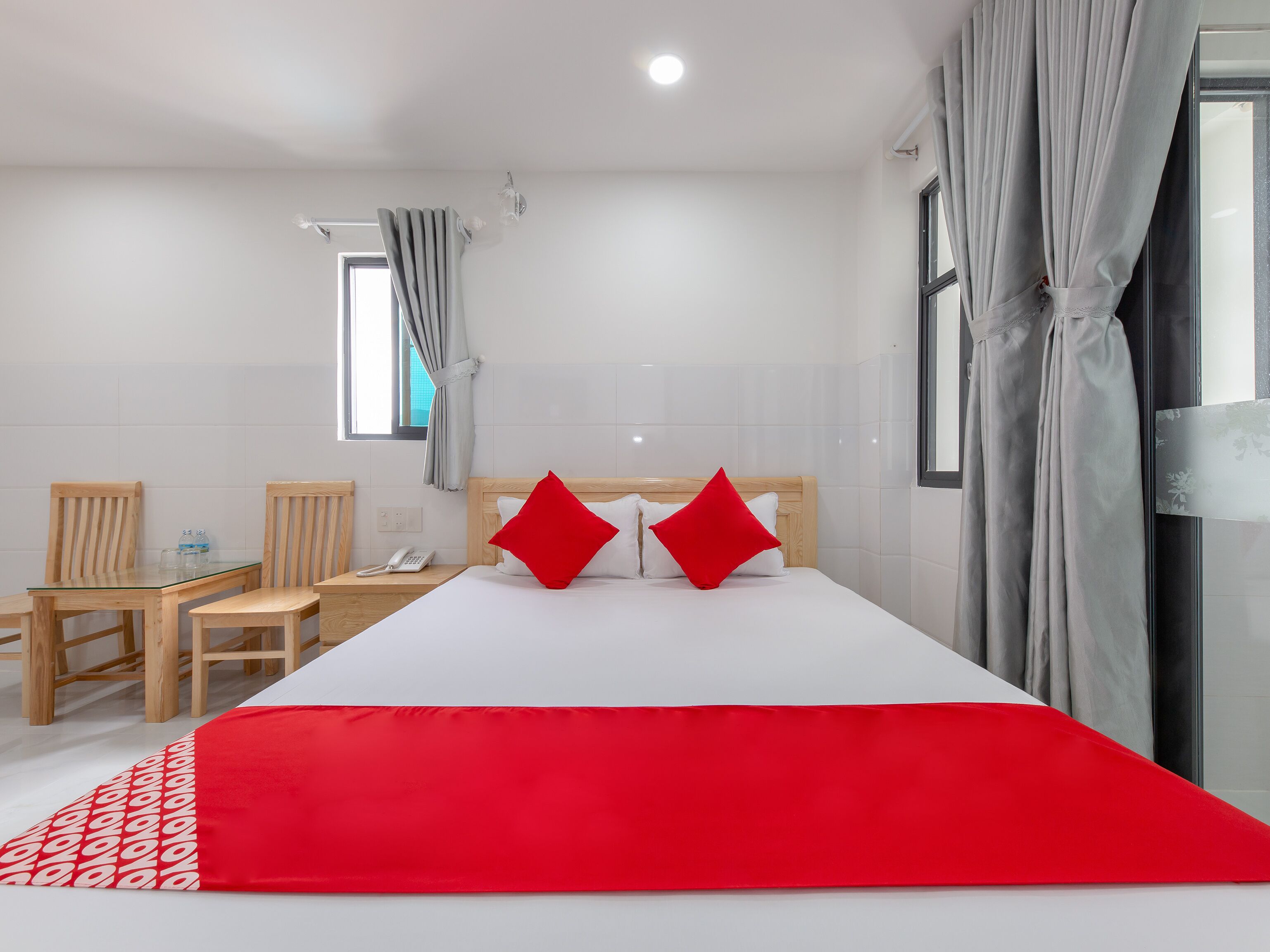 Habitación de hotel con una cama, dos almohadas rojas, una mesita de noche de madera con un teléfono, una mesa de vidrio con sillas y una ventana con cortinas.