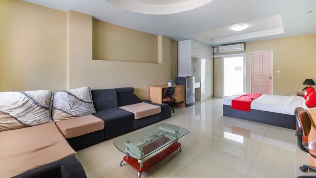 A modern living room with a sofa, a glass coffee table, a bed with a red blanket, a desk with a chair, and a door in the background. - OYO 762 Chompoo (Nakhon Chai Si)