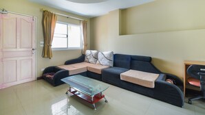 A living room with a sofa, a glass coffee table, a door, and a window with curtains. - OYO 762 Chompoo (Nakhon Chai Si)